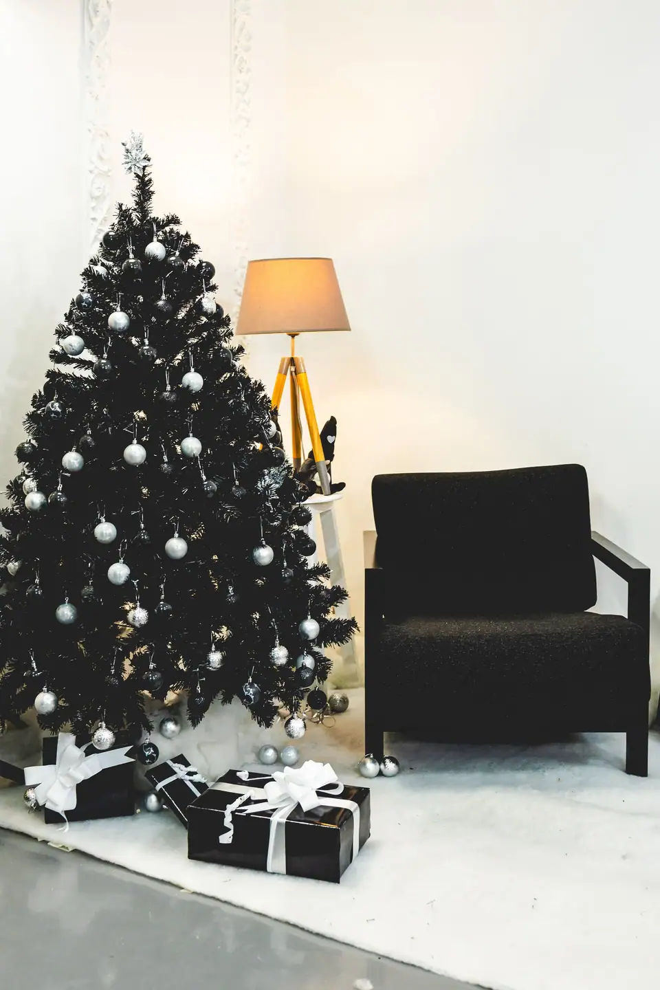 A full black artificial Christmas tree with silver ornaments beside a floor lamp and armchair