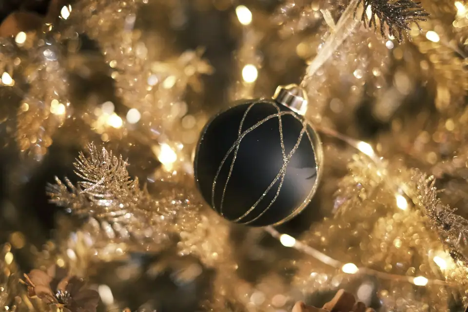A matte black Christmas bauble with gold glitter stripes hanging on a gold tinsel tree with warm white lights