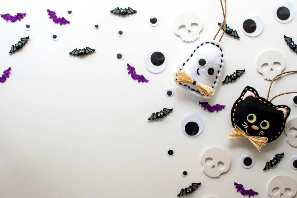 A felt black cat and ghost ornament with small paper bats, skulls and witch hats on a white background
