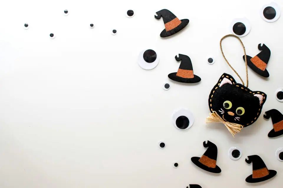 A felt black cat ornament with a hanging loop and raffia bow among small black witch hats on a white surface