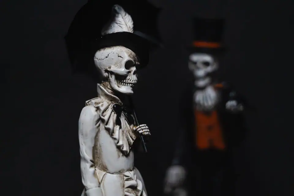 Two skeleton figurines in top hats and ruffled collars against a dark background, the front one holding a black umbrella